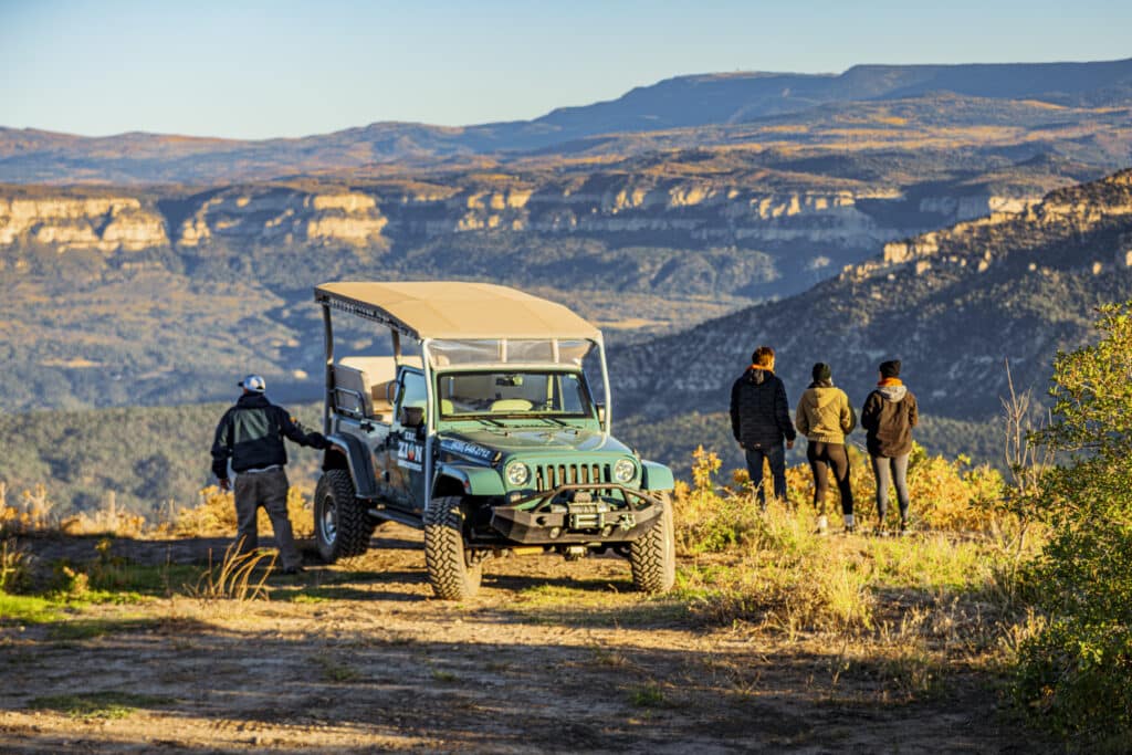 A group of four people stands near a jeep, overlooking a scenic mountain landscape.